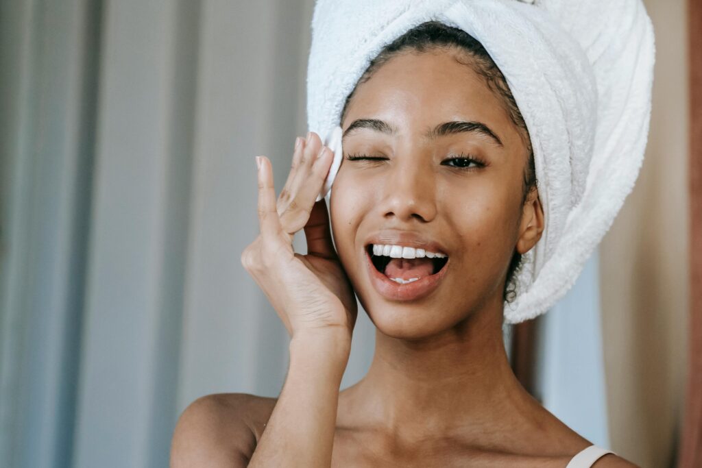 Cheerful young African American female with white towel on head wiping face with cotton pad