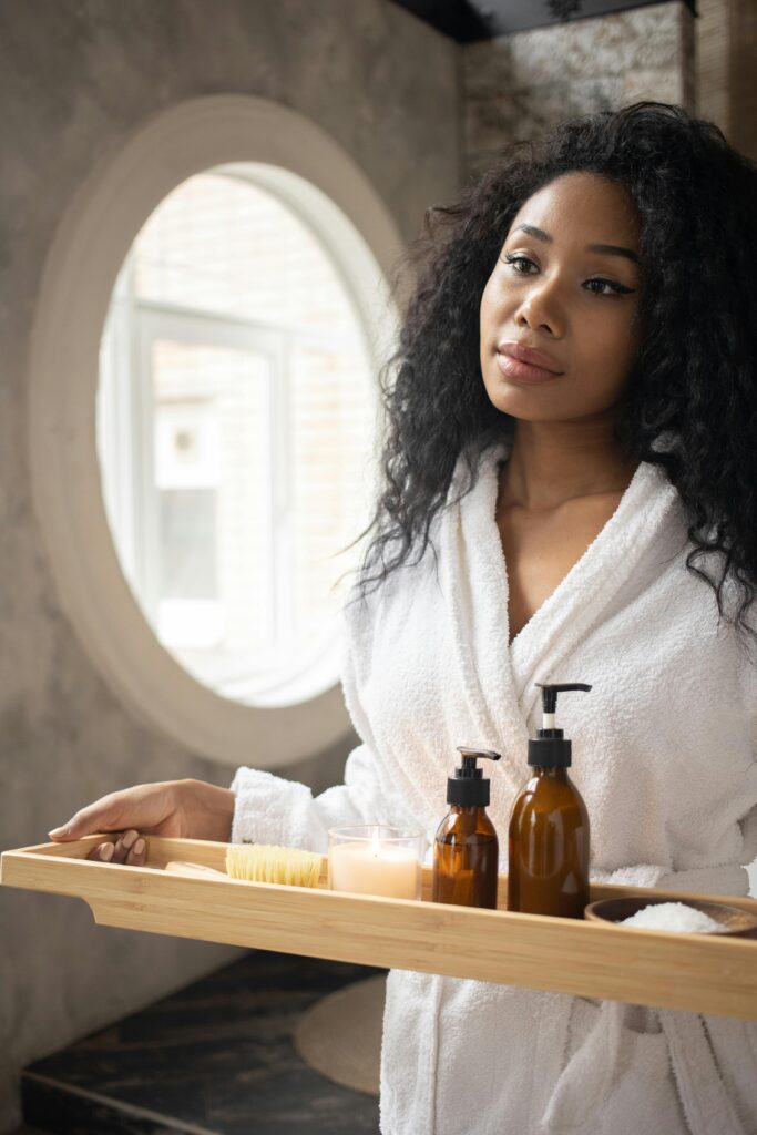 Young ethnic female in white bathrobe holding wooden tray with skin care products standing in bathroom and looking away