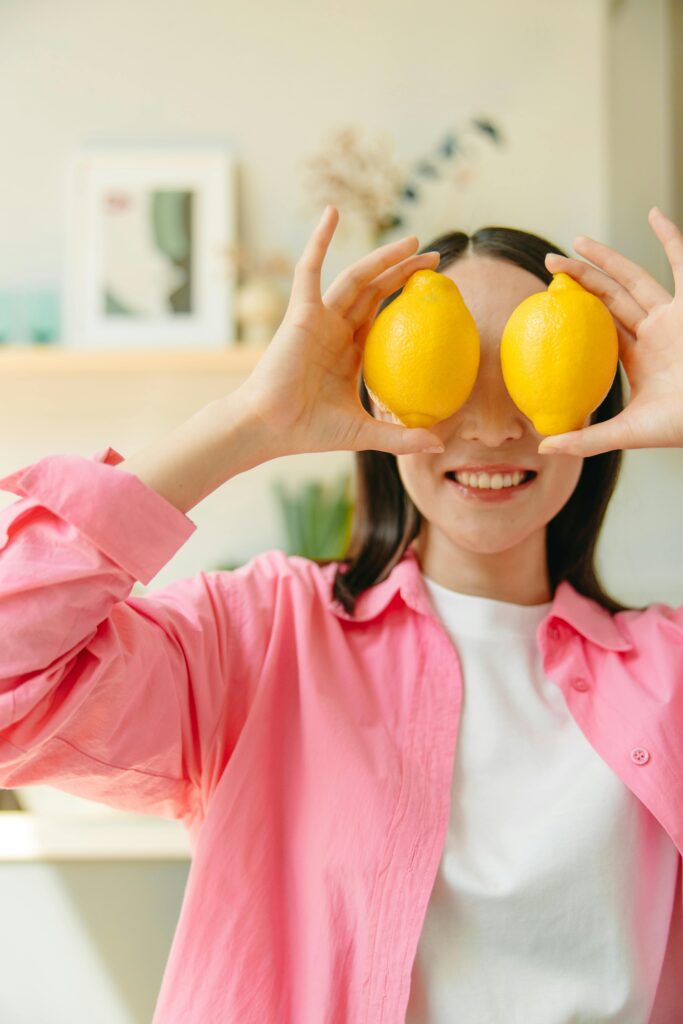 Cheerful woman playfully holding lemons over her eyes indoors.