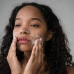 African American female with long dark curly hair washing cheeks with facial foam on gray background
