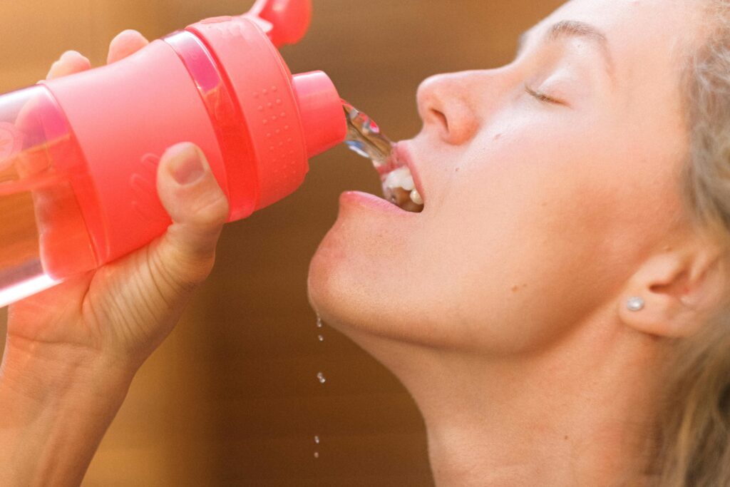 Crop tired young female pouring refreshing clean water into mouth from reusable bottle after intense training