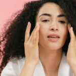 A young woman with curly hair applies skincare products, emphasizing a healthy facial routine.