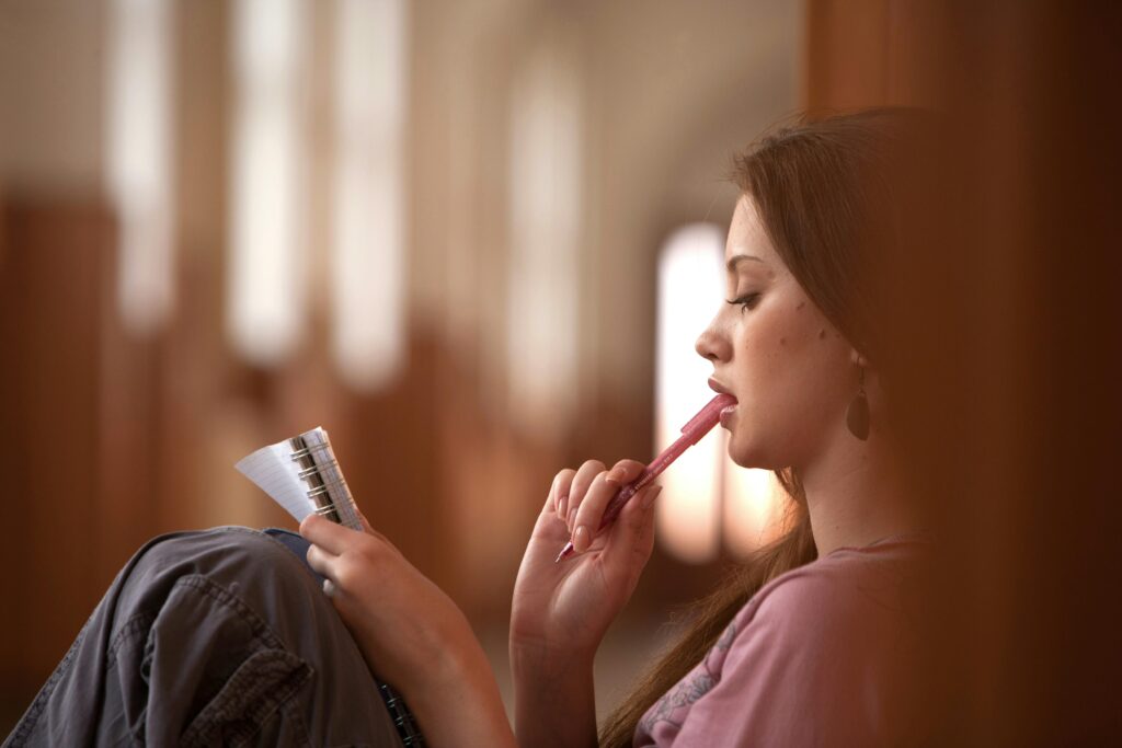 A young woman ponders ideas while holding a pen and notebook indoors, evoking contemplation.