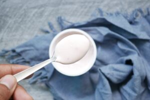 A hand holding a spoon with yogurt above a bowl, placed on a blue cloth.